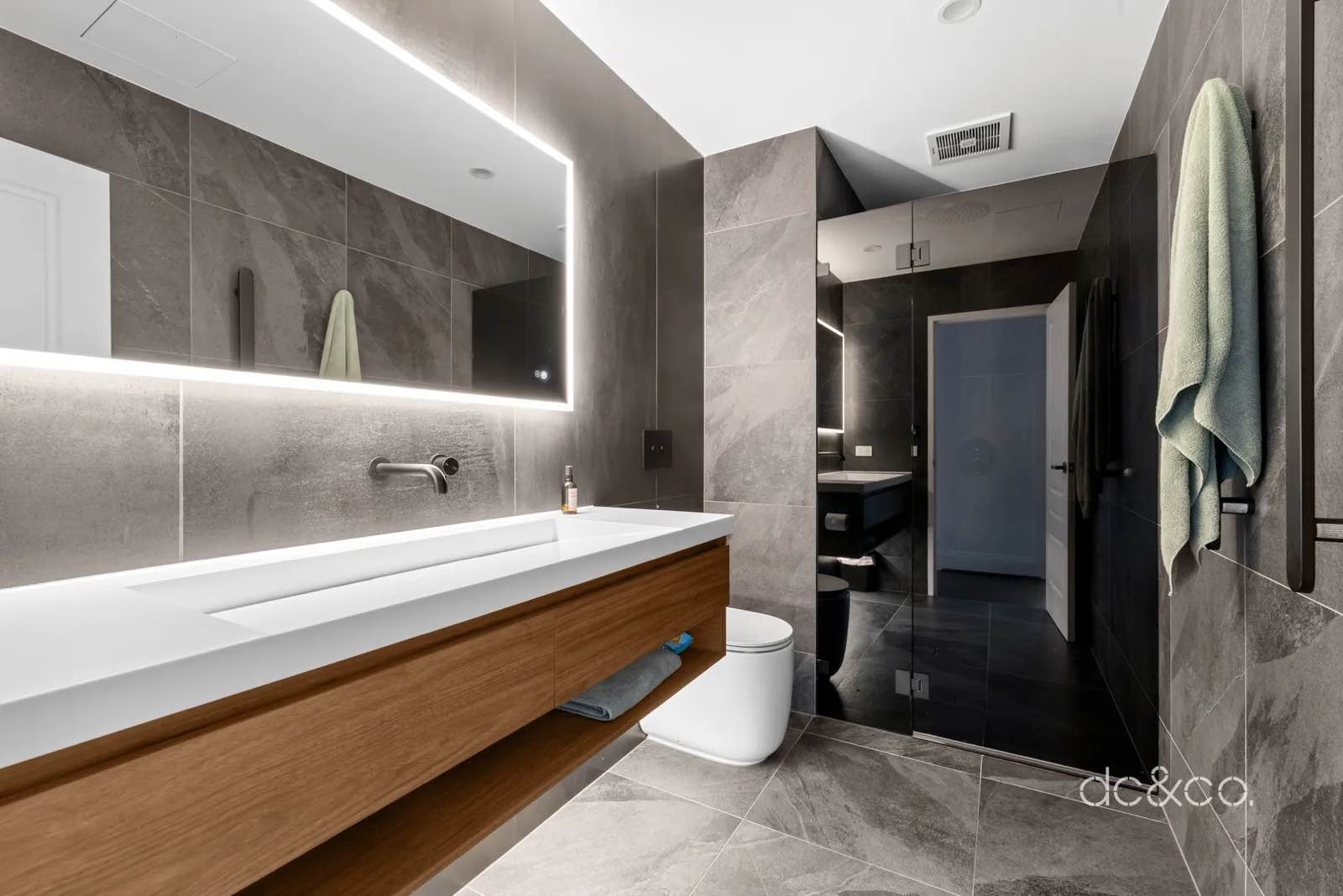 Main bathroom with backlit mirror, timber vanity, trough basin and stone-look tile
